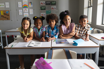 Smiling multiracial elementary school students sitting at desk in classroom - obrazy, fototapety, plakaty