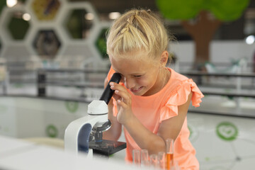 Smiling caucasian elementary schoolgirl looking in microscope while studying in laboratory