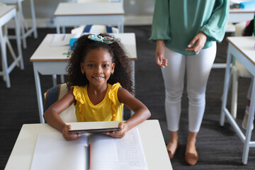 Portrait of smiling african american schoolgirl holding digital tablet at desk in classroom - Powered by Adobe