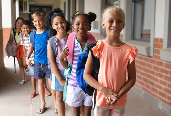 Portrait of smiling multiracial elementary school students with backpack standing in row at school