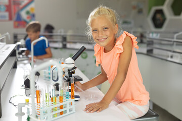 Portrait of smiling caucasian elementary schoolgirl sitting by microscope in school laboratory