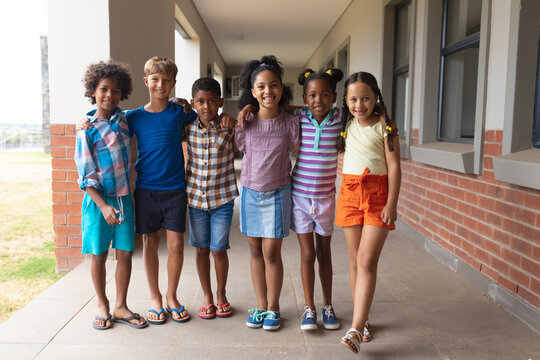 Portrait of smiling multiracial elementary school students with arm around standing in corridor