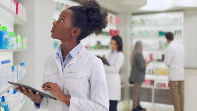 Focused african american woman in white lab coat standing near drugstore showcase and tapping on digital tablet. Competent pharmacist checking expiration date of all medicines.