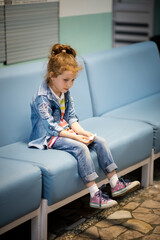 A little sweet girl sits in the lobby of the clinic and waits for an appointment with a doctor. She is wearing denim. She misses it. Health. Medecine. Childhood.