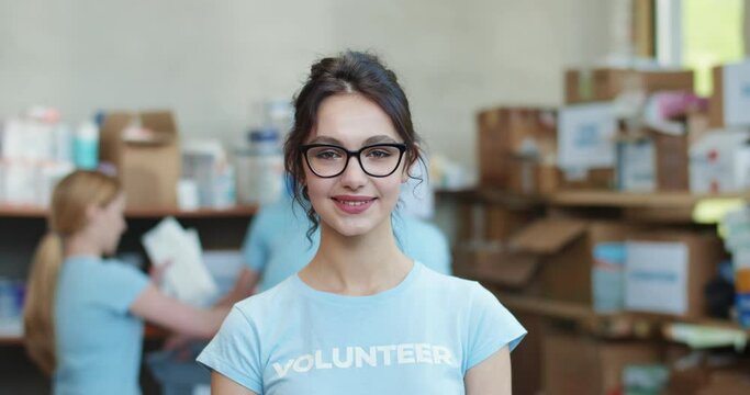 Portrait of attractive young woman in blue volunteer shirt and glasses posing among warehouse of charitable foundation. Group of colleagues working on blur background.