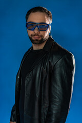 Obraz premium portrait of a man in interactive, cyber glasses. cyberpunk in the studio in black leather clothes. cool trendy handsome guy in flowy leather coat. bearded man with a gun, money on a blue background