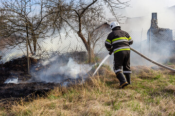 Naklejka premium spring fire, burning dry grass near buildings in the countryside. Firefighter extinguishes the flame. Environmental disaster