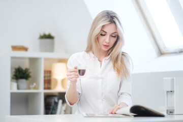 A young beautiful Caucasian blonde woman rests at home. A woman sits at a table, drinking coffee from a glass cup and reading a magazine.