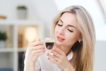 A young beautiful Caucasian blonde woman rests at home. A woman sits at a table, drinking coffee from a glass cup and reading a magazine.