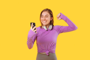 Fototapeta premium Woman wearing sportswear is making the winner gesture while holding phone. Studio shot over yellow background.