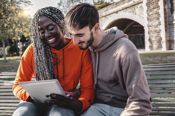 Multi-ethnic couple of young people using digital tablet device browsing content sitting outdoors in the park on a bench - lifestyle concept of biracial couple having fun using tech - Powered by Adobe