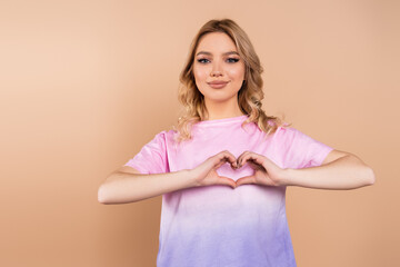 positive woman with wavy hair showing heart symbol with hands isolated on beige.