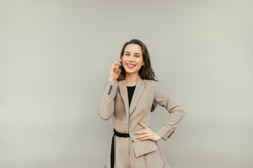 Happy woman with braces on her teeth and in a beige suit communicates on a smartphone with a smile on her face and looks at the camera on a beige background.