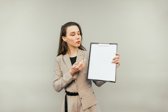 Business woman with a tablet in her hands stands on a beige background looks at a blank white sheet of paper and shows a pen on copy space