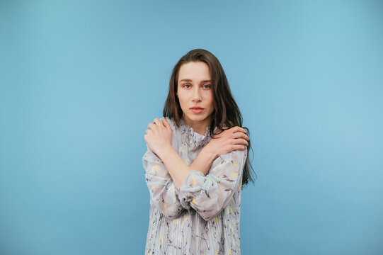 Fragile girl in a dress hugs herself and looks at the camera with a sad face on a blue background