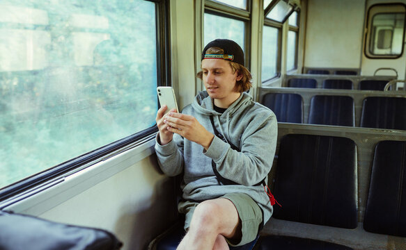 A positive guy with a smartphone in his hands sits in a train by the window while traveling and uses the phone with a smile on his face.