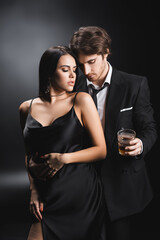 Man in suit holding whiskey near sexy girlfriend in satin dress on black background.