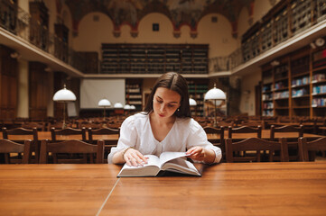 Beautiful woman in a white blouse sitting at a table in a public library and reading a book with a...
