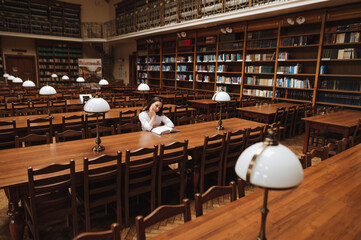 Fototapeta Female student sitting alone in a public old library and reading a book with a serious face. obraz