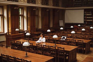 A female student in a blouse sits alone in an empty public library and reads a book.