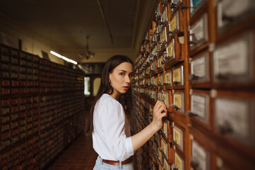 Beautiful woman in a white blouse stands in the archives of the public library, opens a bedside table in search of information and looks at the camera.