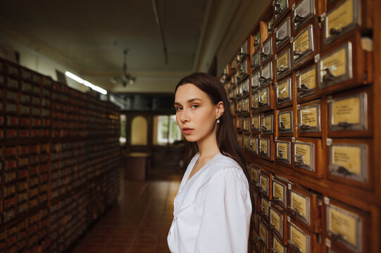 Portrait of a beautiful woman in a white blouse stands in the library archives and poses for the camera with a serious face.