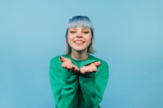 Portrait of a joyful hipster girl with blue hair stands on a blue background with a smile on his face and shows an air kiss.