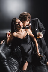 Fototapeta na wymiar Man in suit holding whiskey and kissing sexy woman in silk dress on couch on black background.