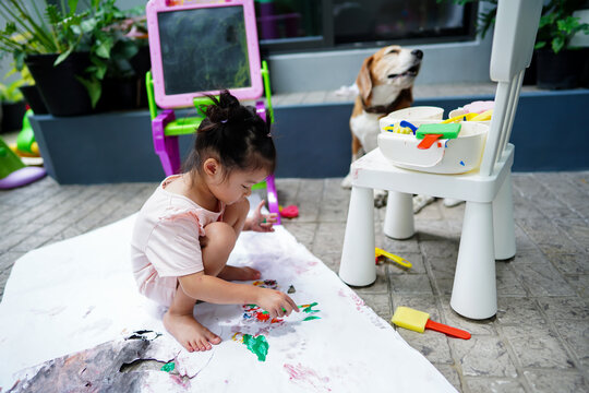 Asian child girl sitting on rip canvas try paintting by paintbrush on rip canvas. Beagle dog near by good her friend.Drawing tools spread all over the tile floor at home garden.Children's creativity.