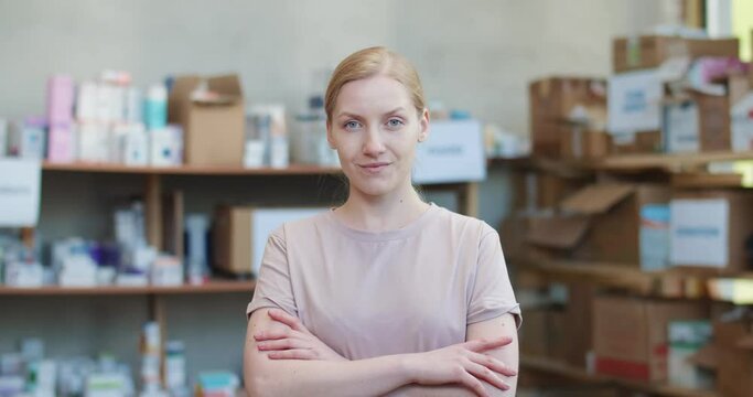 Beautiful caucasian woman looking at camera with serious facial expression and keeping arms crossed while standing at charitable foundation. Blur background of shelves full of food for donation.