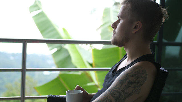 Young man drinking coffee sitting in balcony with exotic garden and sea view. 3840x2160