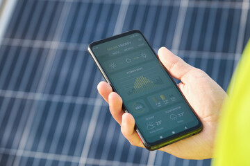 Unrecognizable person showing a solar power generation smartphone app with solar panels in the background. - obrazy, fototapety, plakaty