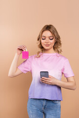 pleased woman holding smartphone and tiny shopping bag isolated on beige.