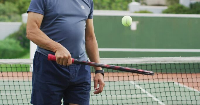 Video of midsection of biracial senior man holding racket and tossing ball on tennis court