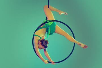 Portrait of young flexible girl, professional air gymnast training on hoop isolated over green...