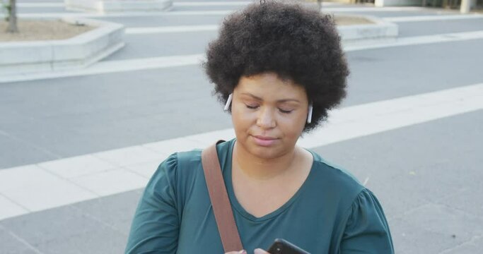 Happy plus size biracial woman using smartphone and wearing earphones in city