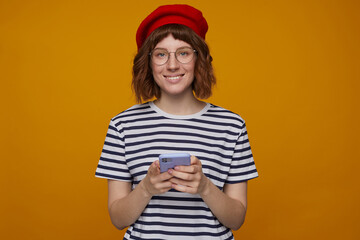 indoor portrait of young ginger female, wears stripped t shirt and glasses posing over orange background looking into camera while holding phone
