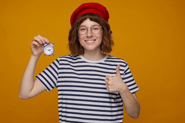indoor portrait of young ginger female, wears stripped t shirt and glasses posing over orange background holding alarm clock and showing thumbs up gesture