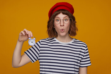 indoor portrait of young ginger female, wears stripped t shirt and glasses posing over orange background holding alarm clock with shocked facial expression