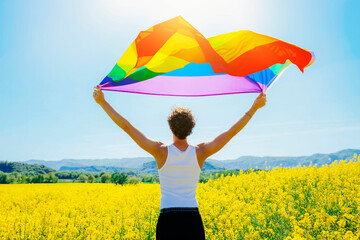 Back view of young man holding gay rainbow pride flag in yellow flowers field. LGBT concept. Spring sunny day celebration.