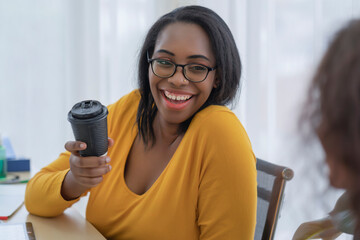 Black woman holding black hot cup of coffee with smiling face while talking with colleague, breaking hour to refresh her body in the afternoon