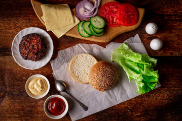 Cooking hamburger or cheeseburger. Different ingredients for a classic hamburger. Grilled meat, vegetables, greens, sauces near a sesame bun. Wooden background.