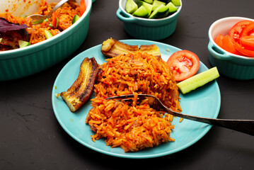 Jollof rice with fried banana on a plate. Rice with tomatoes, onions, spices. Traditional national Nigerian food. Dark background.