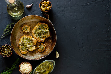 Cauliflower steak with spices lies in a frying pan. Olive oil, chimichurri sauce, herbs, various spices side by side. Dark background. Place for text. Vegetarian food.