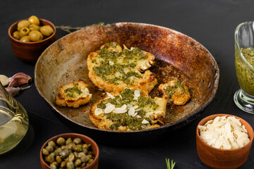 Cauliflower steak with spices lies in a frying pan. Olive oil, chimichurri sauce, capers, olives, herbs, various spices side by side. Dark background. Vegetarian food.