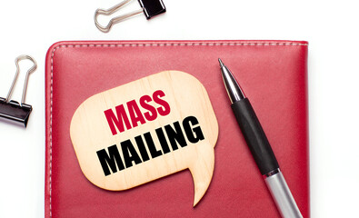 On a light background there are black paper clips, a pen, a burgundy notepad a wooden board with the text MASS MAILING
