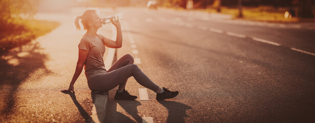 Woman sitting on the border of the roadside after a run and drinking water.