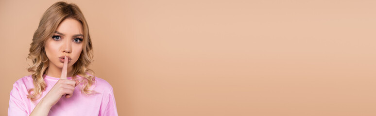 Fototapeta premium serious young woman looking at camera and showing hush gesture isolated on beige, banner.