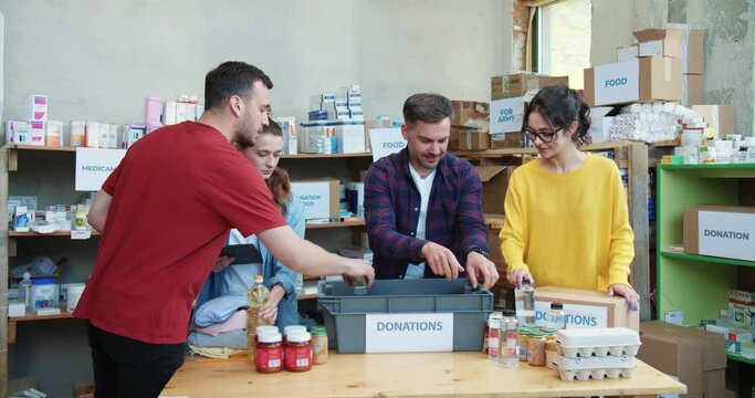 Four young caucasian people dressed in casual clothes using digital tablet and packing donation boxes at warehouse. Helpful volunteers working together for preparing parcels at charity center.