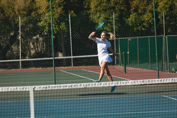 Young female caucasian player jumping while playing tennis game at court on sunny day